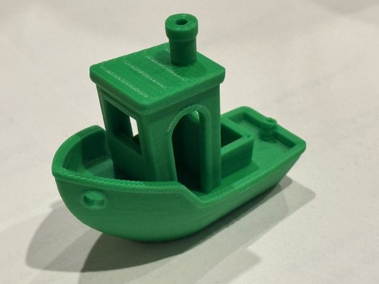 Standard "Benchy" 3D printed model in green PLA against a white background. Benchy is a small stylized cartoon-like boat.