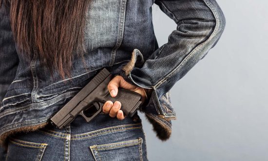 Woman wearing blue denim jacket and pants, viewed from the rear, where we can see she's concealing a semi-automatic handgun.